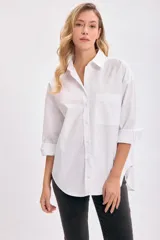 Camisa blanca de manga larga con cuello clásico y botones frontales. Presenta un corte holgado (oversize) y un bolsillo de parche en el pecho izquierdo.
