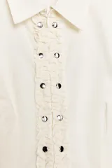 Camisa de manga corta en color crudo, con cuello solapa y cierre frontal mediante cremallera oculta. Presenta un diseño central con volantes verticales y tachas metálicas decorativas a lo largo de la botonadura.