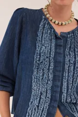 Blusa de jean fino de algodón azul, con escote a la base, mangas abullonadas y puños elásticos. Cuenta con pechera decorada con cintas superpuestas de efecto rústico y desmechado.