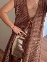 Chaleco largo sin mangas, tejido en punto calado tipo red con hilo de algodón y lurex, color marrón. Presenta escote profundo en V, tiras para atar en el frente, tajo trasero y flecos en el ruedo.