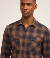 Camisa de franela a cuadros en tonos marrón y azul marino, de corte comfort con mangas largas, cuello inglés, cierre frontal con botones y dos bolsillos con solapa en el pecho.