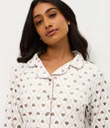 Pijama estilo americano de dos piezas confeccionado en plush. Incluye camisa de manga larga con cuello solapa y cierre frontal de botones, y pantalón largo con cintura elástica y cordón ajustable. Presenta un estampado geométrico de corazones sobre fondo claro.
