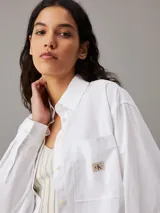 Camisa blanca de manga larga con cuello y botones frontales, corte holgado y bolsillo de parche en el pecho con insignia de monograma.
