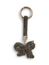 Llavero de metal con acabado en zinc, con un colgante en forma de moño cubierto de pedrería brillante. Incluye una correa de cuero sintético con pedrería y un mosquetón dorado.