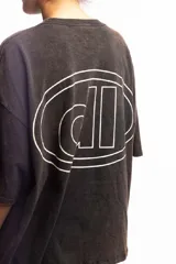 Remera de corte boxy fit, color negro con efecto lavado, de manga corta y cuello redondo. Presenta un logo bordado en el centro del pecho dentro de un óvalo.