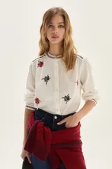 Camisa de algodón color crudo con cuello mao y cierre frontal de botones. Presenta bordados florales en tonos rojo y negro distribuidos en el cuerpo, cuello y puños.