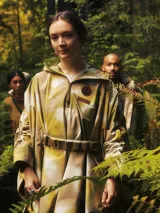 Poncho impermeable de diseño técnico con estampado camuflado en tonos tierra, cuello alto con cierre frontal parcial, capucha integrada y cinturón ajustable con compartimentos. Incluye parches decorativos en el pecho.