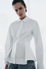 Camisa de popelín de algodón con cuello solapa, manga larga con puños y bajo con diseño peplum. Presenta cierre frontal oculto con botones.
