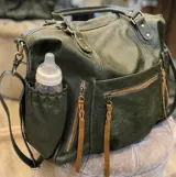Bolso maternal de cuero sintético color verde oliva con textura rugosa. Presenta múltiples bolsillos exteriores con cierres metálicos y tiradores de cuero color suela, además de bolsillos laterales elásticos. Incluye asas superiores y correa ajustable extraíble.