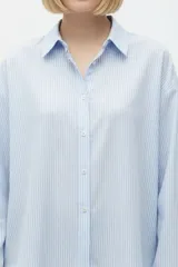 Camisa de algodón a rayas celestes y blancas, de corte holgado, con cuello camisero, cierre frontal de botones y mangas largas con puños rectos.