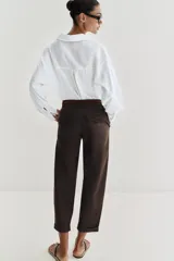 Pantalón chino de tiro bajo color marrón oscuro, con pernera recta y bajo con vuelta. Incluye un cinturón trenzado delgado de color marrón claro. Cuenta con bolsillos delanteros y cierre frontal con cremallera y botón.