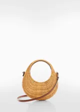 Bolso de mano tipo cesta con estructura circular de ratán natural. Cuenta con un asa corta integrada en la propia estructura y una correa larga ajustable y desmontable de cuero sintético color marrón.