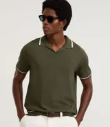 Camisa polo de manga corta color verde oliva, confeccionada en tejido de punto, con cuello y puños con ribetes contrastantes en blanco y rojo.