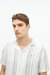 Camisa de manga corta con cuello cubano, confeccionada en una mezcla de lino y algodón con textura. Presenta un diseño a rayas verticales en tonos beige y celeste sobre fondo blanco, con cierre frontal de botones y logo bordado en el pecho.