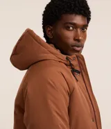 Campera tipo parka color marrón, con capucha ajustable, cierre frontal de cremallera y botones, y múltiples bolsillos con solapa en el frente. Presenta puños ajustables con botones y un diseño funcional de estilo urbano.