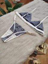 Conjunto de lencería compuesto por un corpiño triangular y una bombacha tipo tanga, confeccionados en encaje gris con detalles de terminaciones en color champagne.
