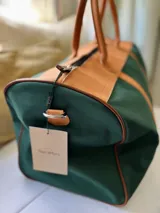 Bolso de viaje tipo duffel de lona negra con detalles en cuero tostado, asas de mano y correa ajustable para el hombro.