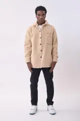 Cazadora de estilo camisa confeccionada en tejido de borreguillo color beige, con cierre frontal de botones, cuello clásico y bolsillo de parche en el pecho.