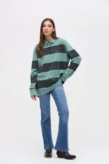 Remera tipo polo de manga larga con diseño de rayas horizontales anchas en bloques de color verde y negro. Presenta cuello clásico, cierre frontal con botones y un pequeño bordado en el pecho. Confeccionada en algodón lavado con textura suave y corte relajado.