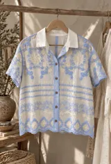 Camisa blanca de manga corta con bordados en tonos celestes y crudos, cuello camisero liso, cierre frontal con botones y diseño holgado.