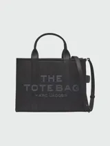 Cartera tipo tote de cuero granulado color tostado, con diseño estructurado y rectangular. Presenta doble asa superior fija y una correa larga ajustable y extraíble. En el frente, destaca el logo estampado con la inscripción 'THE TOTE BAG MARC JACOBS'.