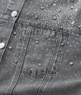 Campera de jean gris con efecto desteñido, corte oversized, cuello inglés, cierre frontal con botones niquelados y aplicaciones de brillos y piedras en la parte superior delantera.