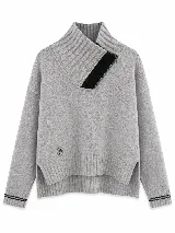 Polera tejida en lambswool color gris, con cuello alto y cierre frontal parcial con detalle en negro. Presenta diseño de tajos laterales en el ruedo, puños acanalados con detalle de rayas en contraste y un pequeño aplique circular con logo en el frente.