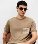 Remera Marfinno de manga corta y cuello redondo, de color tostado, confeccionada en tejido de punto con mezcla de poliéster, elastano y poliamida. Cuenta con un bolsillo frontal aplicado en el pecho con diseño geométrico en color blanco.