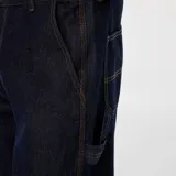 Pantalón de jean de mezclilla oscura, corte recto y holgado, con costuras contrastantes en color mostaza/amarillo. Presenta un bolsillo utilitario lateral en la pierna derecha, característico del estilo carpintero.