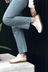 Zapatilla de estilo urbano en tonos marfil, con detalles en color dorado en el talón y ojales metálicos. Presenta suela de goma color caramelo y cordones al tono.