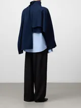 Cazadora corta de gabardina en color azul marino, con cuello chimenea y cierre frontal de botones. Presenta un diseño estructurado con mangas amplias y una pieza de tela integrada que cae lateralmente a modo de capa o chal.
