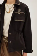 Campera estilo militar o safari color verde oliva, confeccionada en algodón. Presenta cuello solapa y bolsillos frontales con tapa. Destaca por sus apliques de pedrería o bordados en tonos marrones y dorados en el cuello y sobre los bolsillos.