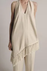 Top halter color crudo, de escote en V y diseño confeccionado en hilatura con mezcla de viscosa. Presenta bajo asimétrico con flecos al tono y cierre en la espalda.