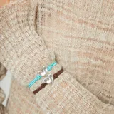Pulsera de hilo elástico doble color turquesa, con dije central de corazón plateado de forma irregular y un cierre de bola metálico plateado.