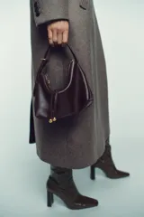 Bolso de mano tipo saca, color borgoña, con asa de mano ajustable y correa bandolera extraíble. Presenta un detalle de doble tira con terminaciones de bolas metálicas doradas en el frente.