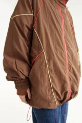 Campera rompevientos beige de nylon con diseño oversize, cuello alto, cierre frontal negro y detalles de costuras y ribetes en contraste.