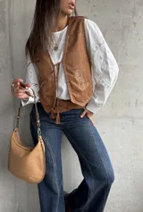 Chaleco de gamuza color camel con bordados decorativos en hilo claro y cierre frontal mediante lazos con borlas. Presenta un diseño de inspiración boho con terminaciones delicadas.