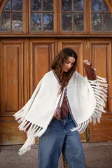 Poncho tejido de color blanco, con flecos en los bordes y detalles de bordado en crochet a lo largo de la abertura frontal. Cuenta con un cierre de cordones con borlas en el cuello.