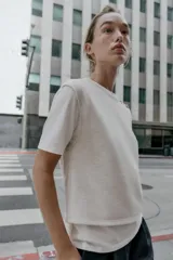 Top de cuello redondo y manga corta con diseño de capas que combina una remera de algodón blanca con un chaleco de punto gris superpuesto. Presenta detalles de botones joya en el hombro.