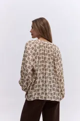 Blusa de corte relajado con estampado geométrico en tonos beige y negro. Presenta cuello en V con detalle de frunces, mangas largas abullonadas con puños elásticos y una caída fluida.