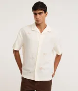 Camisa masculina boxy de manga corta en tricolina color marfil, con cuello cubano, cierre frontal con botones y sutiles bordados veraniegos en tono tostado.