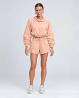 Conjunto deportivo de dos piezas color durazno, compuesto por un buzo cropped de manga larga con cuello alto ajustable y cintura regulable, y un short con cintura clochard y cordones laterales.