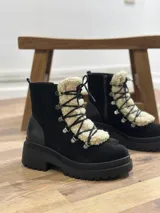 Bota tipo borcego de cuero marrón con suela track negra de goma. Presenta un aplique frontal desmontable de corderito sintético color crema y cordones negros con ganchos metálicos.