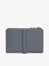 Billetera bifold Tommy Hilfiger de diseño compacto en color gris. Presenta un cierre superior con cremallera y un detalle frontal decorativo con broche metálico que incluye el logo de la marca. Cuenta con múltiples compartimentos internos para tarjetas y billetes.