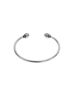 Pulsera tipo brazalete abierto de metal plateado con terminaciones en forma de esfera en los extremos.