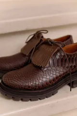 Mocasín de cuero vacuno negro con textura tipo reptil, suela track de goma, cordones finos y solapa con flecos sobre el empeine.