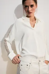 Camisa blanca de manga larga con cuello solapa, cierre frontal con botones y bajo asimétrico. Presenta detalles de puntilla combinada a tono en las mangas y hombros.