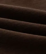 Pantalón de sarga color marrón oscuro, con corte super baggy, cintura con trabillas, bolsillos laterales y traseros, y cierre frontal con botón y cremallera. Incluye una cadena metálica decorativa en la cintura.