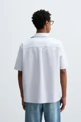 Camisa de algodón con rayas verticales finas azules y blancas, corte relajado (relaxed fit), cuello bowling y manga corta. Posee un bolsillo plastrón en el pecho y aberturas laterales en el bajo.