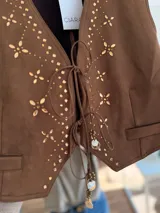 Chaleco de gamuza sintética color marrón, con escote en V y cierre frontal mediante lazos ajustables. Presenta detalles decorativos de tachas metálicas y apliques brillantes en el frente, además de dijes colgantes en los lazos.